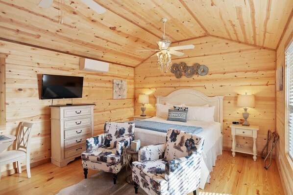 Ein gemütliches Schlafzimmer in einer Blockhütte mit Bett, zwei Nachttischen, einer Kommode, einem Fernseher und floralen Ohrensesseln.
