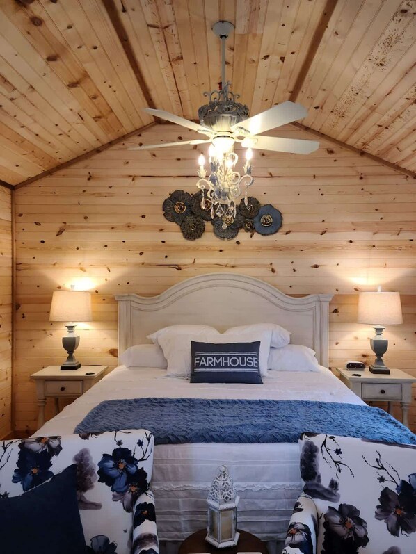 Un dormitorio acogedor con techo de madera, una araña, una cama con almohada 'FARMHOUSE', dos mesitas de noche con lámparas y un farol decorativo.