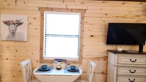 Interior de una cabaña de madera con una mesa de comedor para dos, un televisor en un tocador y una ventana con persianas.