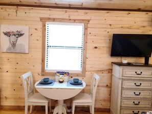 A wooden cabin interior with a dining table set for two, a television on a dresser, and a window with blinds.