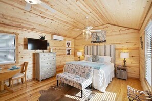 Een knus slaapkamer in blokhutstijl met een houten plafond, een bed met witte beddengoed, een dressoir, een televisie en een bureau met stoel.