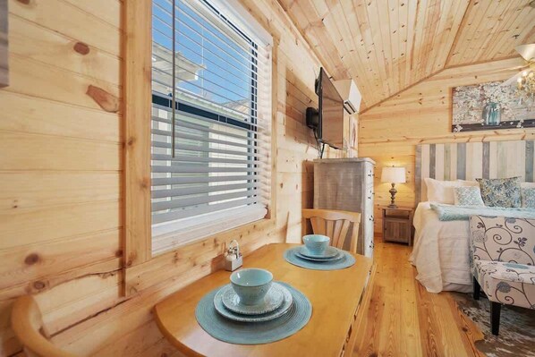 A wooden cabin interior with a dining table, chairs, a bed, and a television mounted on the wall.