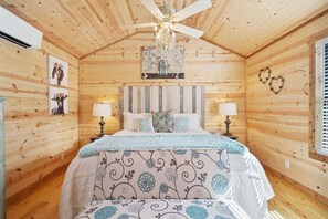 A wooden cabin bedroom with a bed, two bedside lamps, a chandelier, and wall decorations. - Romantic Cabin Right on Main St! (Fredericksburg)