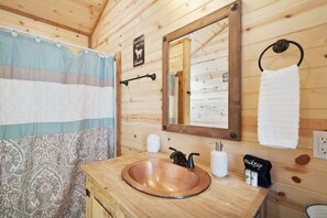 A wooden cabin bathroom with a copper sink, a mirror, a towel ring, and a shower curtain. - Romantic Cabin Right on Main St! (Fredericksburg)
