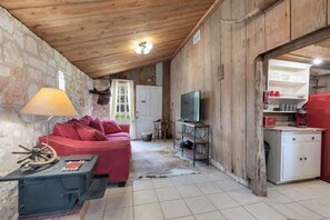 A rustic interior with a stone wall, wooden ceiling, a red sofa, a television, and a small kitchenette.