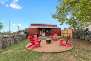 Ein rustikales Bauernhaus mit roter Fassade, einem Holzzaun und einer Feuerstelle mit roten Adirondack-Stühlen im Garten.