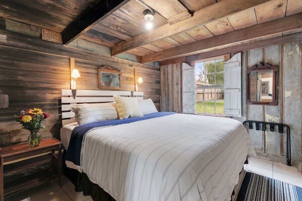 Ein rustikales Schlafzimmer aus Holz mit einem großen Bett, zwei Nachttischlampen, einem Nachttisch mit Blumenvase und einem Fenster mit Fensterläden.