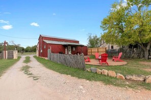 Une grange rouge, avec un chemin de gravier menant à l’entrée, entourée d’une clôture en bois et agrémentée d’une petite terrasse avec des chaises rouges.