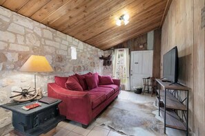 Ein gemütlicher Raum mit einem roten Sofa, einer Holzbalkendecke, einer Steinwand, einem Fernseher und einem Tisch mit einer Lampe.