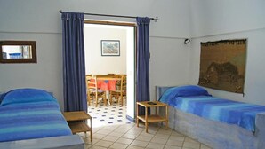 Habitació amb dos llits, una taula petita, una cadira i vista a una zona de dinar a través d'una porta.