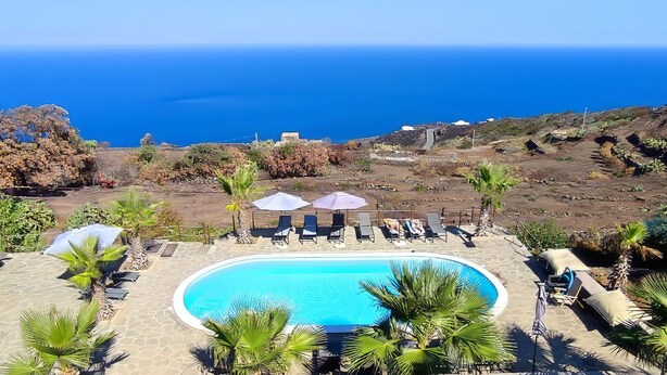 Zona de piscina amb gandules i ombrel·les, envoltada de vegetació i amb vista a un paisatge costaner.
