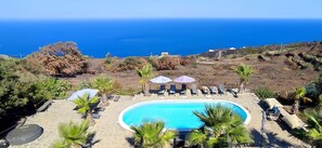 A pool area with lounge chairs and umbrellas, surrounded by greenery and overlooking a coastal landscape. - Simple Cozy family apartment in villa with pool (Pantelleria)