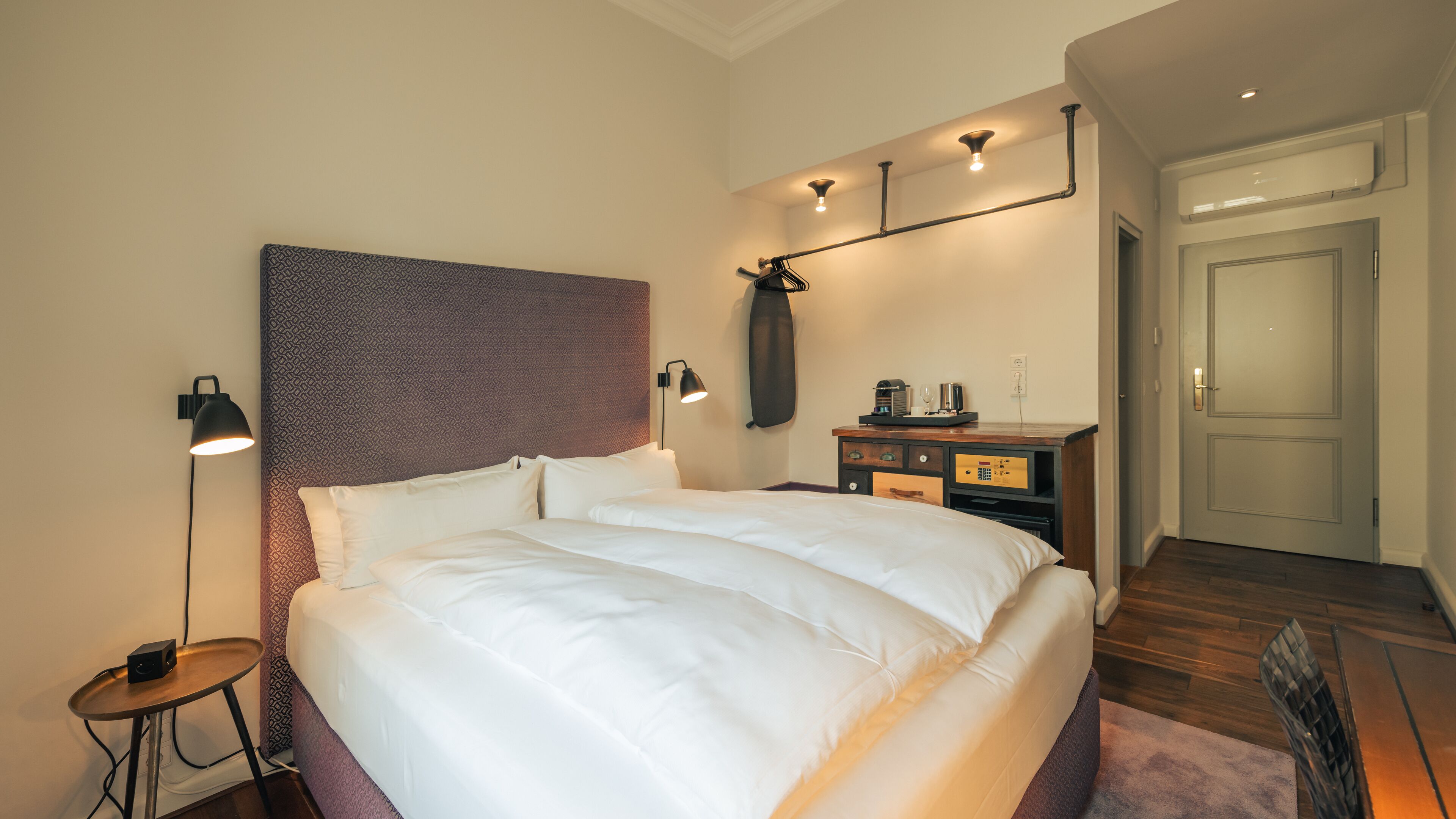 Una habitación de hotel ordenada con una cama grande, lámparas de noche, una mesita con un altavoz, un armario y una puerta.