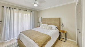 A bedroom with a bed, a bedside table, a lamp, and a window with curtains. - Charming condo close distance to Honeymoon Island! (Dunedin)