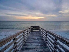 A wooden pier leading to a beach with a calm sea and a sunset in the background.