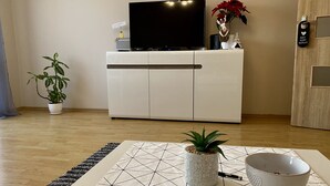 A modern living room with a flat-screen TV on a white cabinet, a small potted plant, a decorative vase with red flowers, and a wooden door with a key lock. - Cosy Apartament in Rzeszów City Centre (Rzeszów)