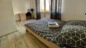 A bedroom with a bed, a desk, a chair, a standing fan, and a window with curtains. - Cosy Apartament in Rzeszów City Centre (Rzeszów)