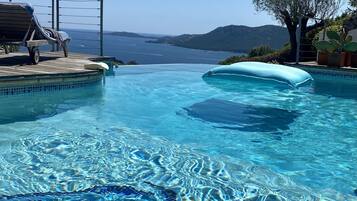 Une piscine aux eaux cristallines, offrant une vue sur la mer et les montagnes, entourée de chaises longues et d’une terrasse en bois.