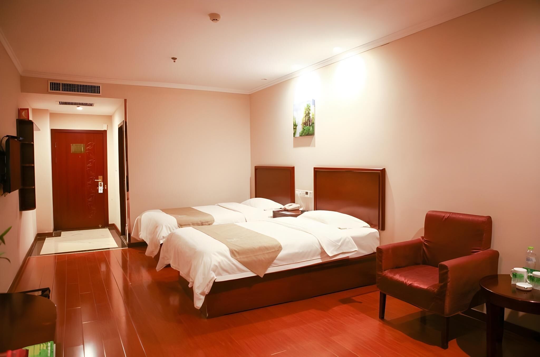 A hotel room with a double bed, a red armchair, a small table with a bottle and a cup, and a wall-mounted TV.
