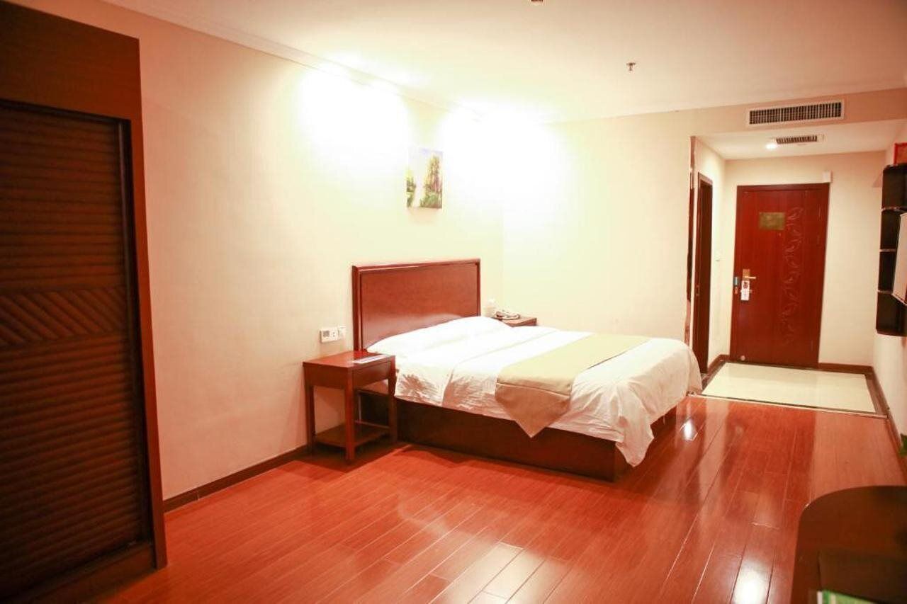 A hotel room with a wooden floor, a bed with white bedding, a nightstand, a wall-mounted TV, and a door with a red handle.