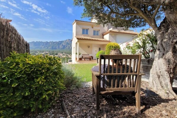 A wooden bench in a garden with a view of a house and mountains in the background.
