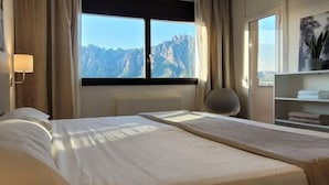 A hotel room with two beds, a window with a mountain view, a chair, and a shelf with towels.