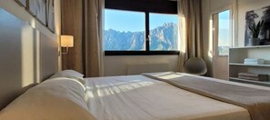 A hotel room with two beds, a window with a mountain view, a chair, and a shelf with towels.