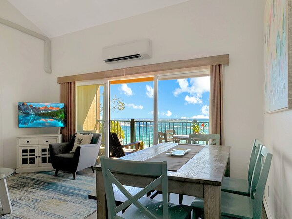 A room with a large window overlooking a scenic view, a dining table with chairs, a sofa, a TV, and a cabinet. - Updated Beachfront Condo! Walk to Old Town Kapa'a (Kapaa)