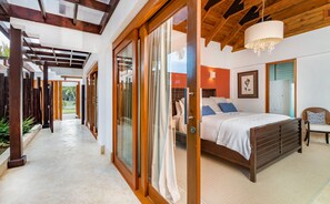 A bedroom with a large bed, a bedside table, and a chair. There is a window with curtains, a ceiling fan, and a wall-mounted light fixture.