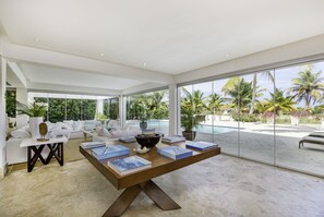 A spacious living area with a large wooden table, white sofas, and a view of a swimming pool and palm trees.