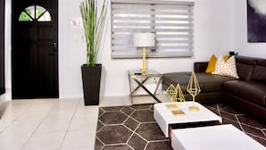 Een woonkamer met een zwarte deur, een potplant, een glazen tafel met een lamp, een bruine leren bank en een geometrisch vloerkleed.