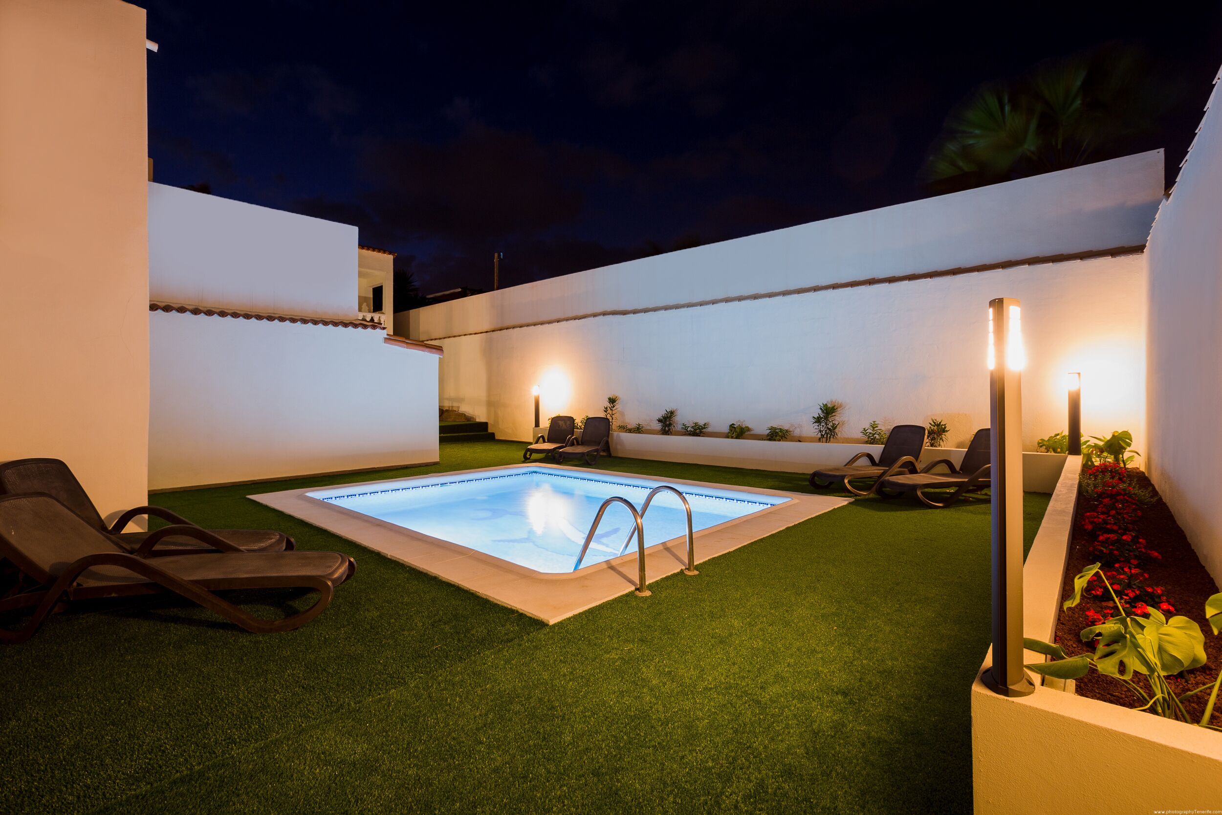 Una piscina con una pequeña zona de terraza, rodeada de césped artificial y sillas de descanso.