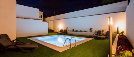 Una piscina con una pequeña zona de terraza, rodeada de césped artificial y sillas de descanso.