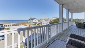 Eine überdachte Veranda mit weißen Geländern, Korbmöbeln und Blick auf den Strand und Häuser im Hintergrund.