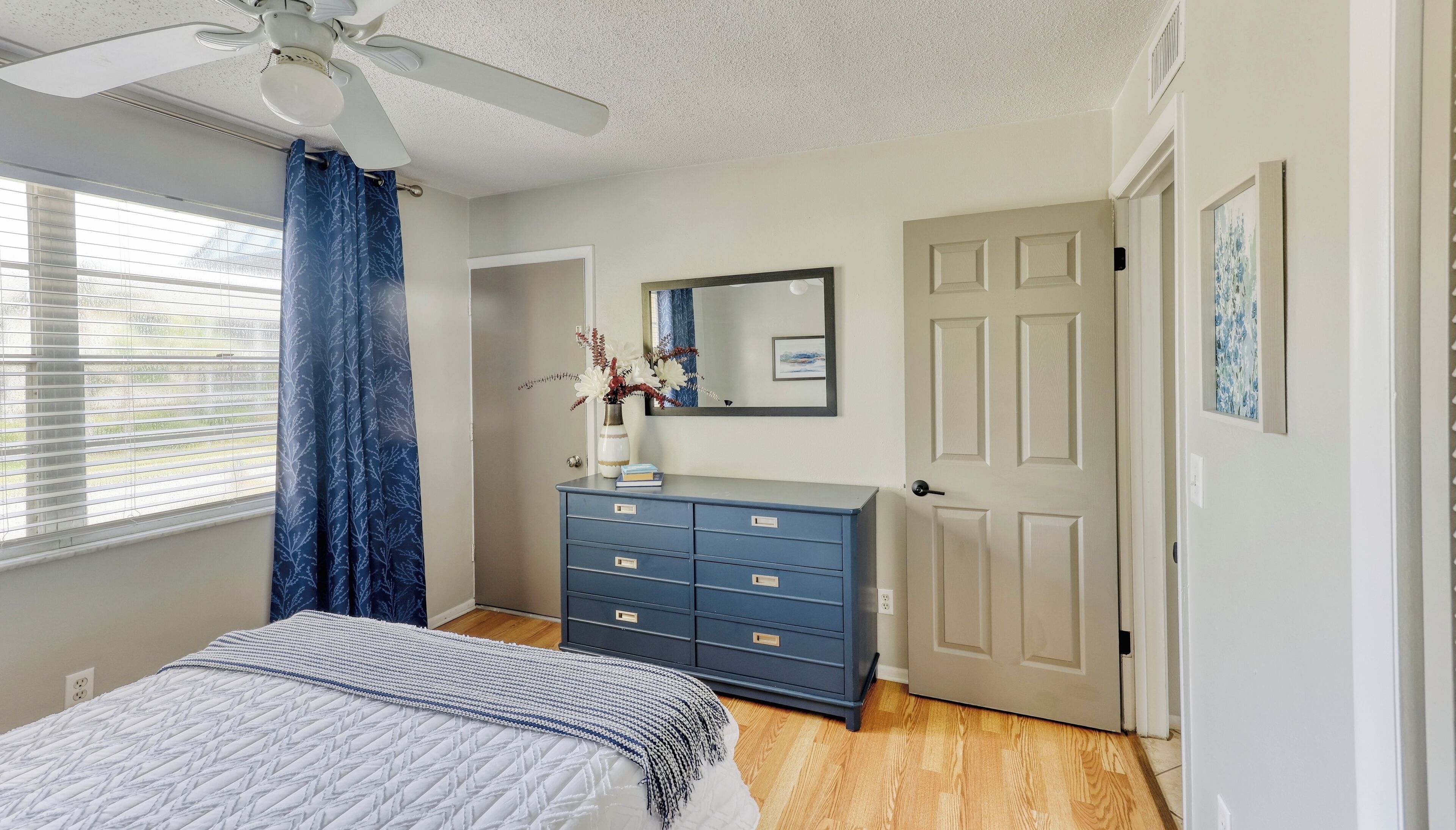 A bedroom with a bed, a dresser, a ceiling fan, a door, and a window with curtains.