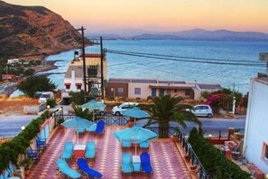 A coastal property with a terrace featuring sun loungers and umbrellas, overlooking a body of water and a mountainous landscape. - Adonis Hotel (Agios Vasileios)