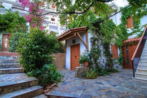 A stone-paved courtyard with steps leading to a wooden door, surrounded by lush greenery and a pergola. - Adonis Hotel (Agios Vasileios)