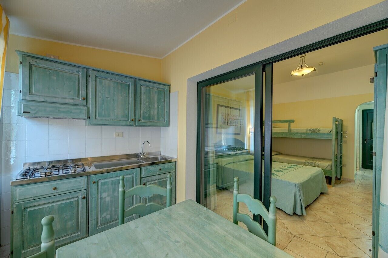 Une cuisine compacte équipée d’une cuisinière à gaz, d’un évier et de meubles de rangement verts. Une table à manger avec des chaises se trouve au premier plan. Une porte-fenêtre coulissante donne accès à une chambre où se trouve un lit superposé.