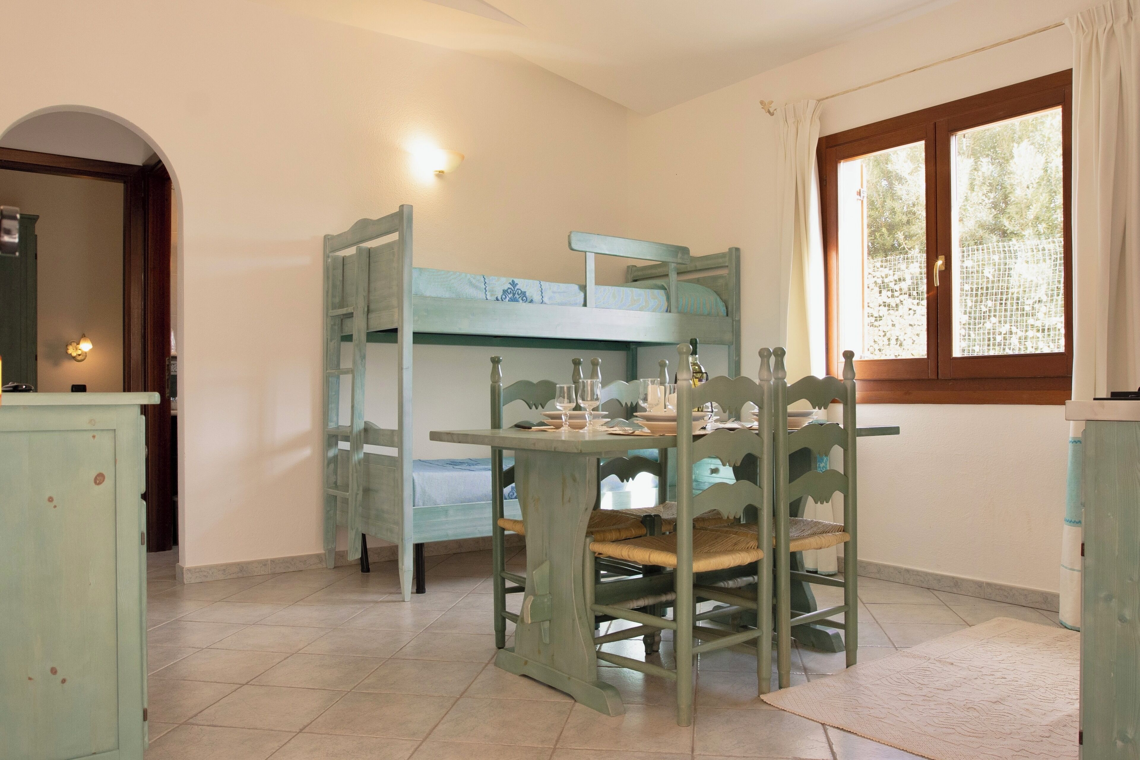 A room with a bunk bed, a dining table with chairs, a window with curtains, and a door leading to another room.