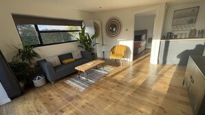 A modern living room with a sofa, coffee table, and a yellow chair. There is a large window and a plant in the corner. - Maygrove Retreat - Sleeps 2 - Jacuzzi & Boat Hire (Staines)