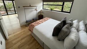A bedroom with a bed, pillows, a bedside table, a hanging rack, and a window with a view of greenery. - Maygrove Retreat - Sleeps 2 - Jacuzzi & Boat Hire (Staines)