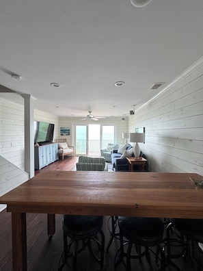 Un vaste espace de vie comprenant une table à manger en bois, des tabourets de bar, un téléviseur et une vue sur l’extérieur.