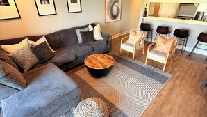 A modern living room with a grey sectional sofa, a round coffee table, and a ceiling fan. There are framed pictures on the wall and a kitchen area in the background. - Waterfront Walden Lodge! Discounted Monthly Stays! (Montgomery)