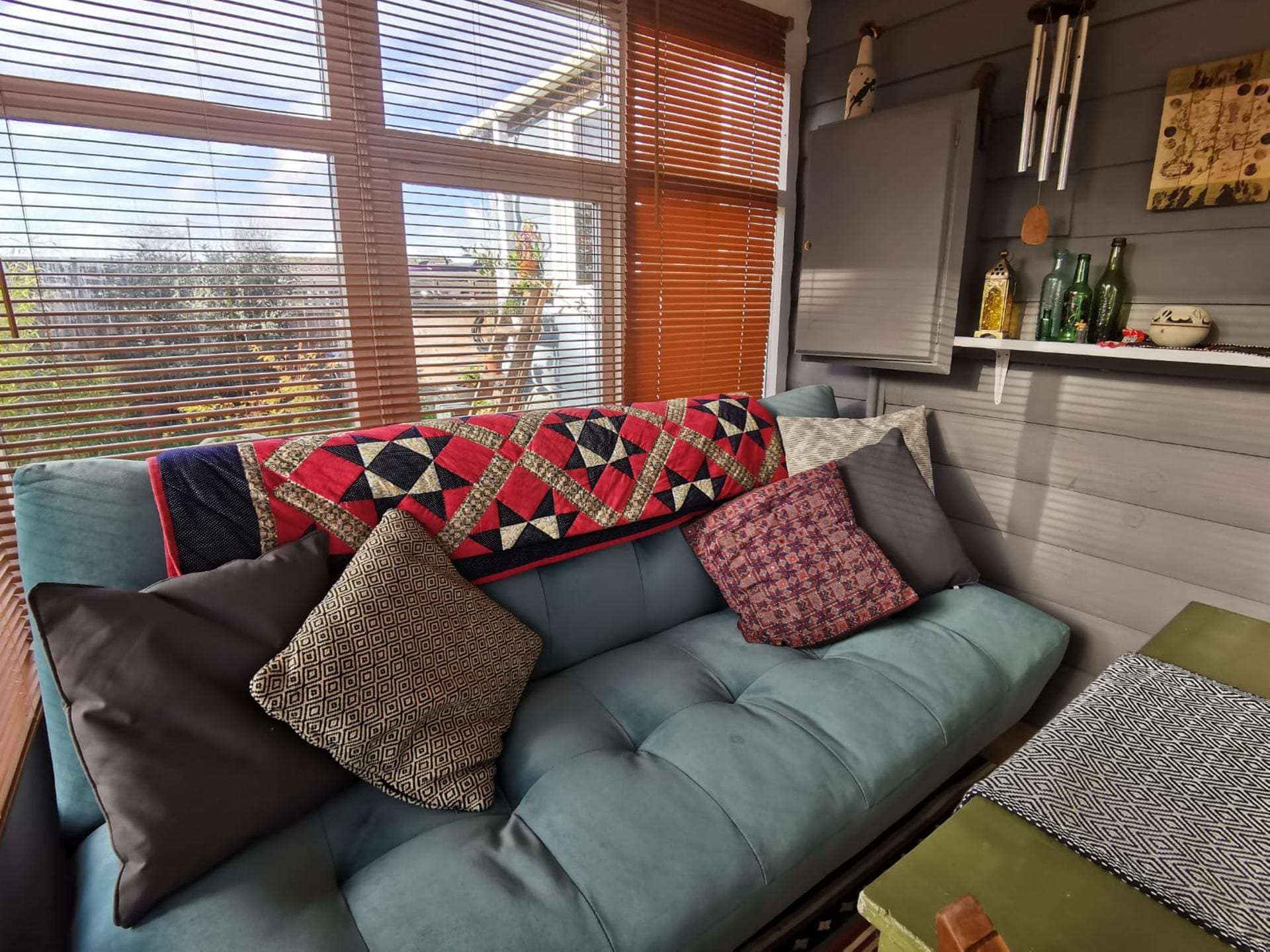 A blue sofa with patterned cushions and a quilt, a small table with a patterned table runner, and a wall with a shelf holding decorative items.