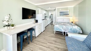 A compact room with a bed, a desk, a chair, a TV, and a small kitchen area.
