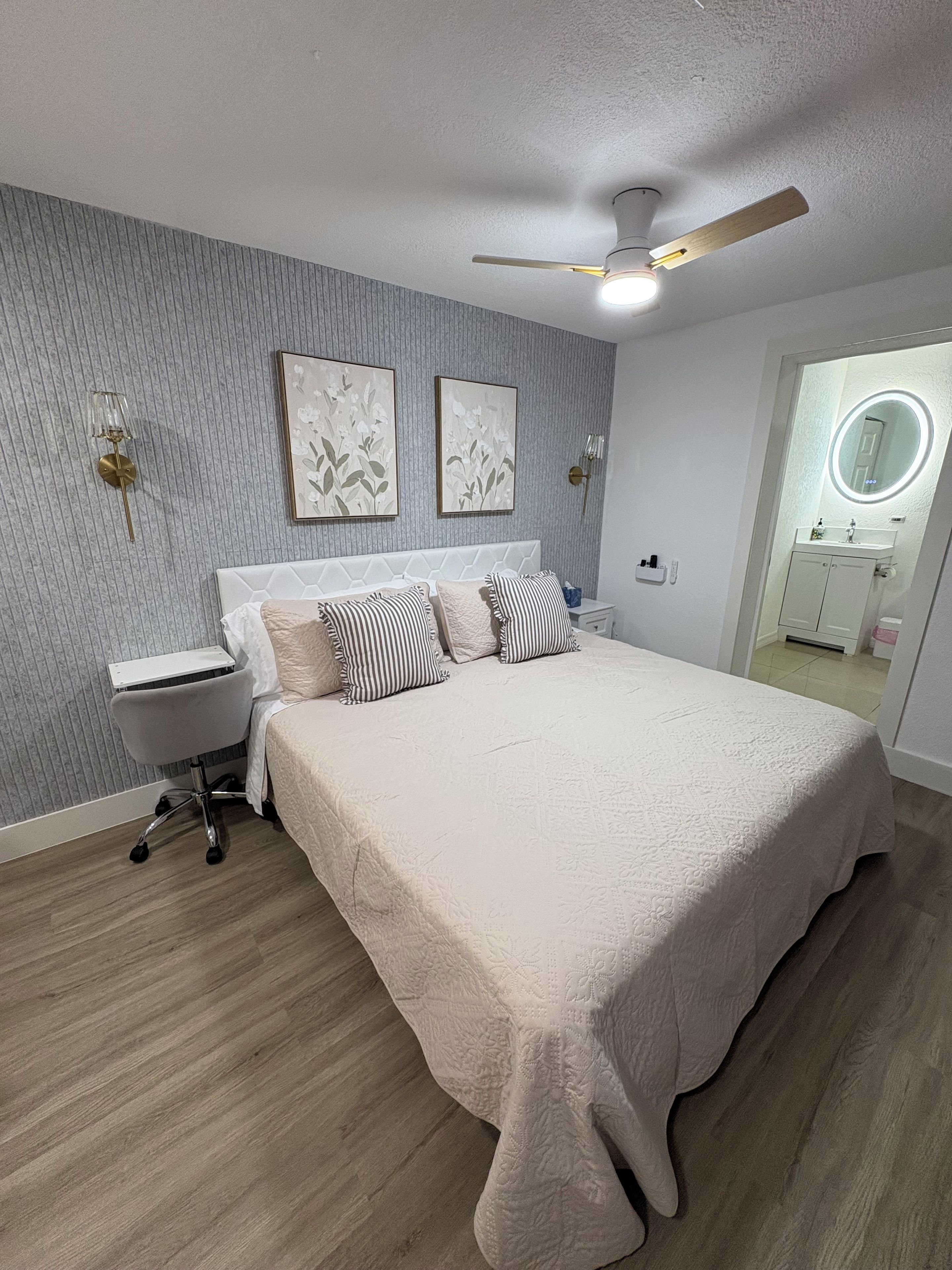 A bedroom with a bed, a bedside table, a chair, a ceiling fan, and a wall-mounted light fixture.