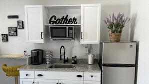 A kitchen with white cabinets, a microwave, a coffee maker, a sink, and a refrigerator. There are decorative signs on the wall and a plant on the counter. - little paradise (Tampa)