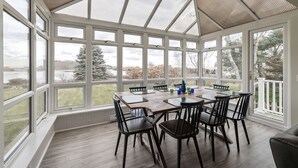 Ein Wintergarten mit Holztisch und -stühlen, großen Fenstern und Blick auf Bäume und ein Gewässer.
