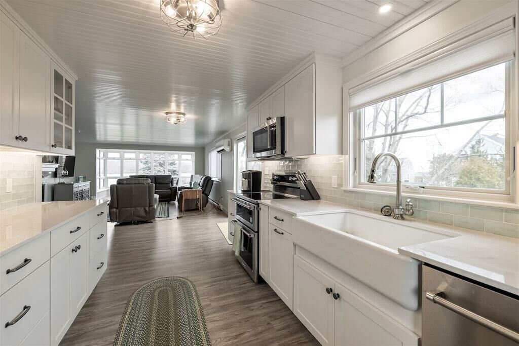 A modern kitchen with white cabinetry, a large window, and a dining area in the background.