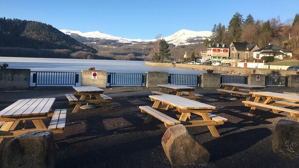 Un área de picnic junto al lago con bancos y mesas de madera, un cartel de prohibido estacionar y vistas de una montaña y edificios al fondo.